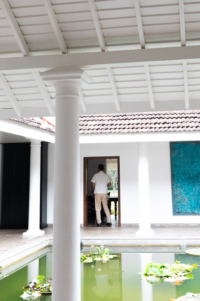 Uga Riva: A New Boutique Hotel In Negombo With A 180-Year History 5 A member of staff dressed in a white linen shirt and chinos walking through a door of a courtyard. In front there is a pond with lily pads and a white pillar is blurred out.