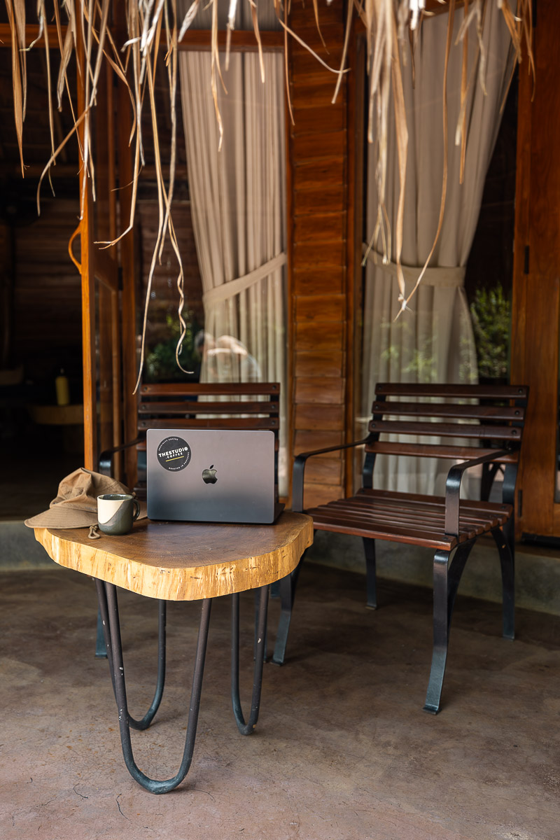 Gal Oya Lodge: A Luxury Eco Lodge In Sri Lanka's Least Visited National Park 18 A small wooden table and wooden chairs outside a bungalow at Gal Oya Lodge eco-hotel in Sri Lanka. There is a laptop on the table with a TheStudio.Coffee sticker, as well as a safari hat and a cup of coffee.