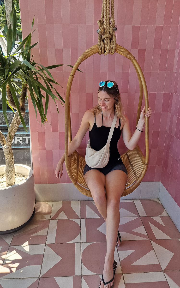 Vegan Canggu: A tried-and-tested guide to vegan cafes & restaurants in Bali's digital nomad hotspot 8 Jodie sitting on a swing outside KYND Vegan Cafe in Canggu, Bali. The wall behind is pastel pink and it is sunny. The tiles on the ground are branded with the KYND logo.